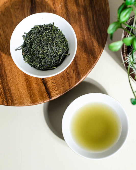 Overhead view of loose-leaf green tea and a cup of brewed green tea in white bowls on a wooden tray with plant decor.